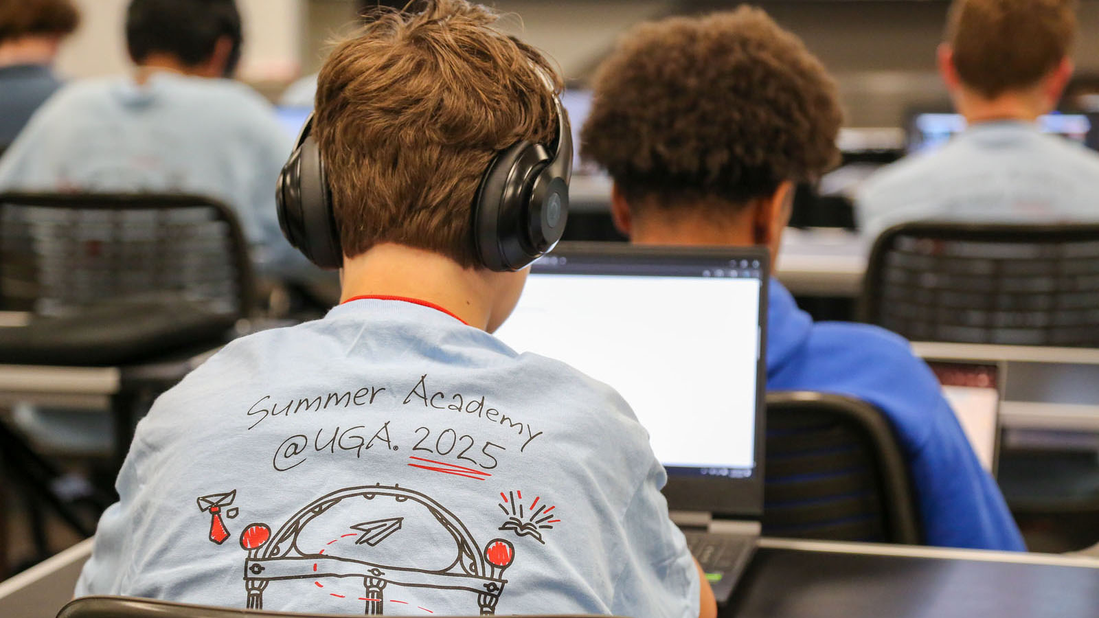A student sits at a computer and "Summer Academy at UGA 2025" is on the back of his shirt.